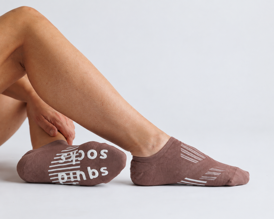 Person seated in brown no-show grip socks, with one sole turned toward the camera. By Squid Socks.
