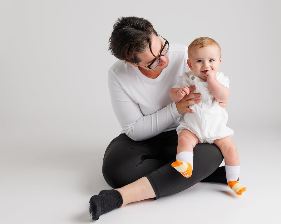 Adult seated beside a baby wearing Candy Corn grip socks.