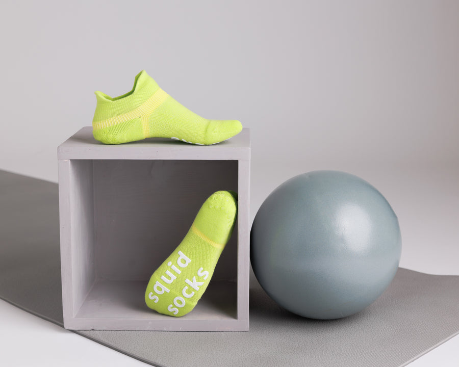 Lime green ankle grip socks displayed on a cube beside an exercise ball, showing side and sole views. By Squid Socks.