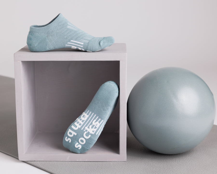 Teal no-show grip socks displayed on a cube beside an exercise ball, showing top and sole views. By Squid Socks.