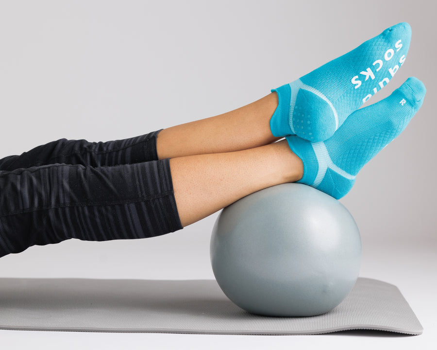 Person wearing turquoise ankle grip socks with feet resting on an exercise ball. By Squid Socks.