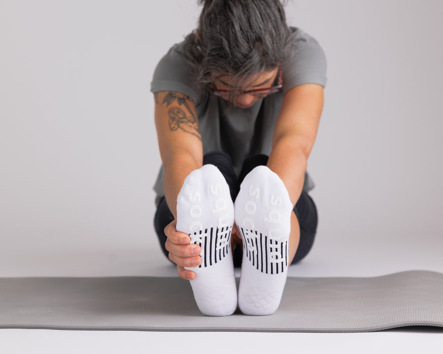Person stretching forward on an exercise mat in white no-show socks, showing black bottom grips. By Squid Socks.