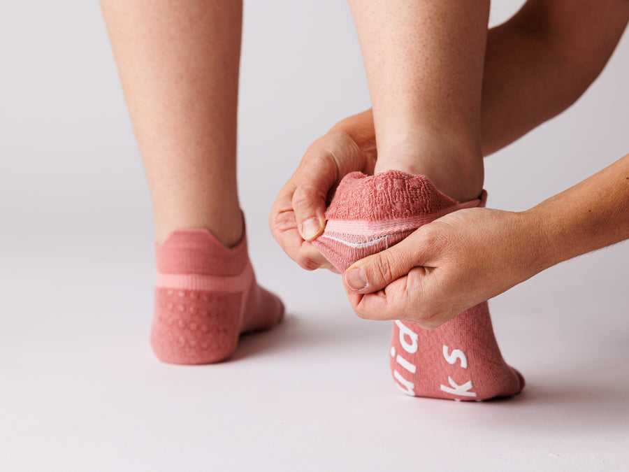 Person pulling back a coral ankle sock at the heel, with white bottom grips visible. By Squid Socks.