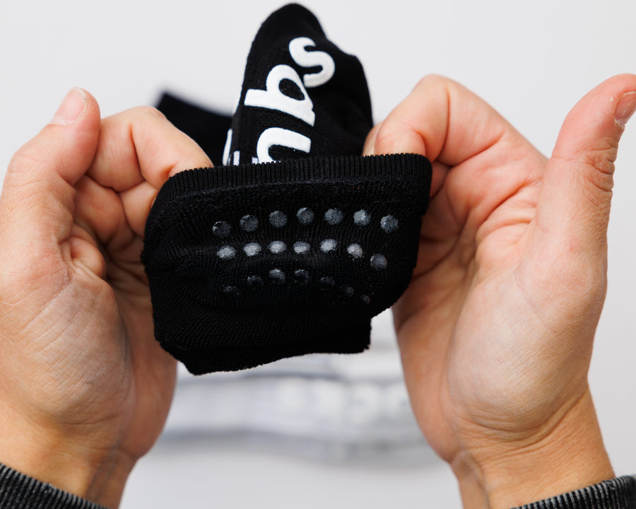 Hands holding open a black no-show sock to show grip dots inside the heel. By Squid Socks.