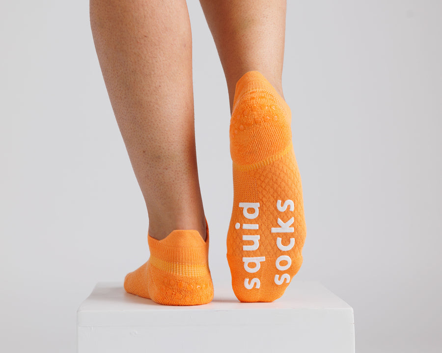 Orange ankle grip socks viewed from behind, with one sole raised to show white bottom grips. By Squid Socks.