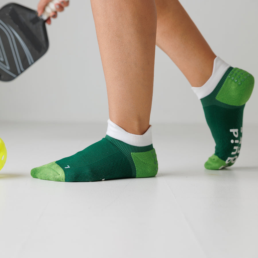Green ankle pickleball grip socks worn beside a pickleball paddle, with one heel raised. By Squid Socks.