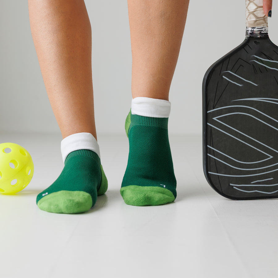 Green ankle pickleball grip socks worn beside a pickleball and paddle, showing lime toe panels. By Squid Socks.