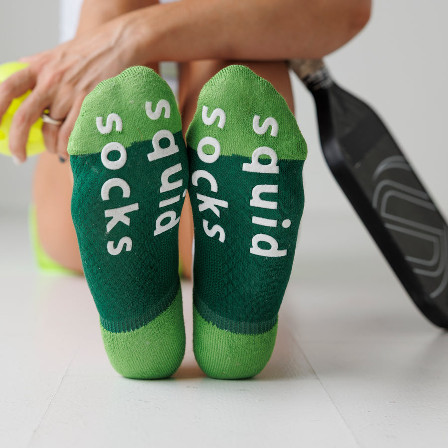 Green Pickle Pack ankle socks viewed from the soles, showing white bottom grips beside pickleball gear. By Squid Socks.