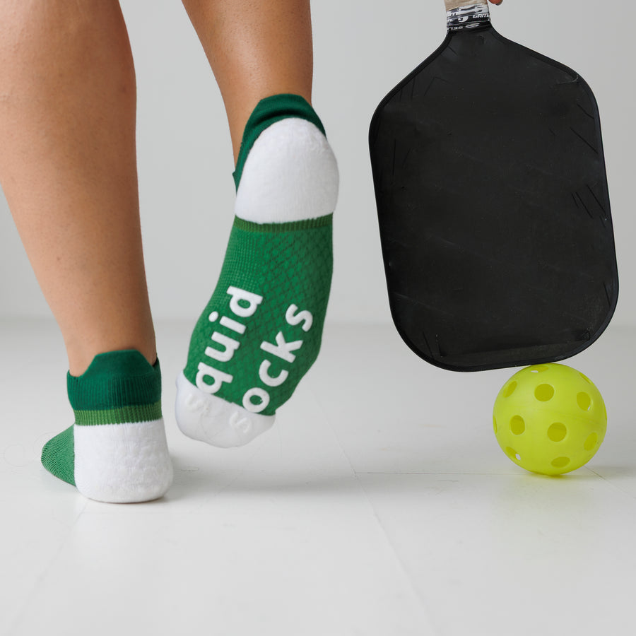 Green and white ankle pickleball grip socks with one sole raised beside a pickleball paddle and ball. By Squid Socks.