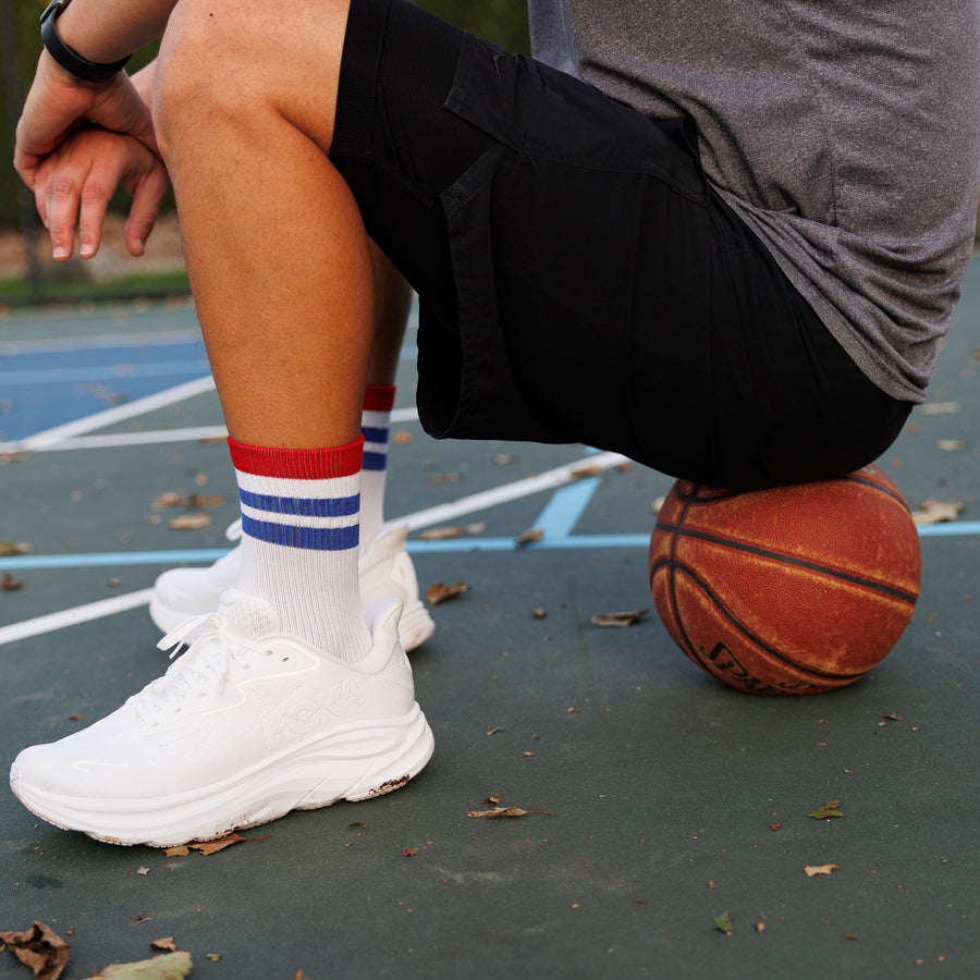 Person wearing white, red, and blue striped crew socks and white sneakers beside a basketball on a court. By Squid Socks.