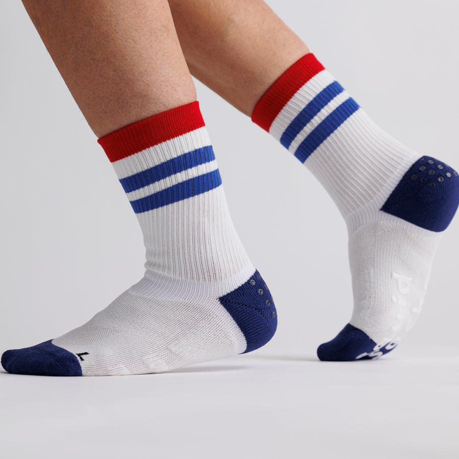 White All-American crew grip socks with red cuffs and blue stripes worn with one heel raised. By Squid Socks.