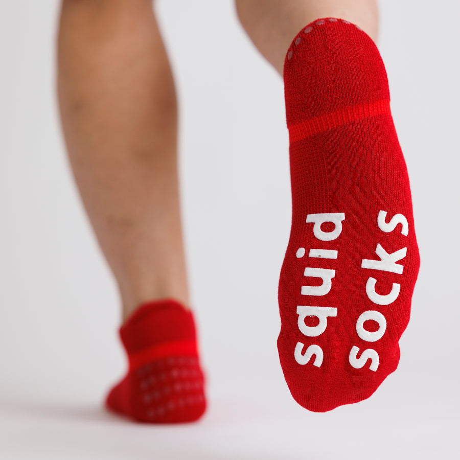 Red ankle grip socks with one foot lifted to show the white bottom grips. By Squid Socks.
