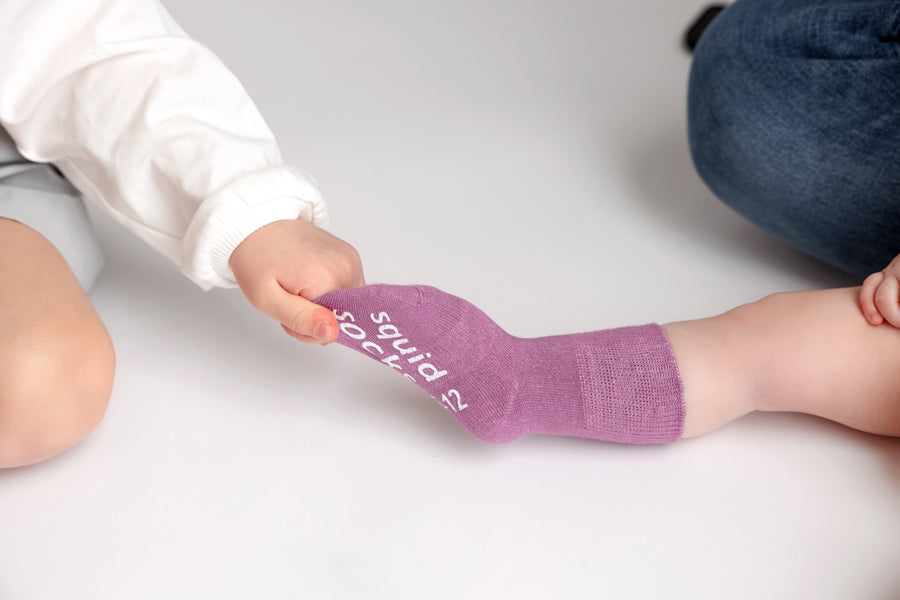 Orchid kids socks: child gently pulling a bright lavender sock on a baby. Squid Socks, the brand seen on Shark Tank.