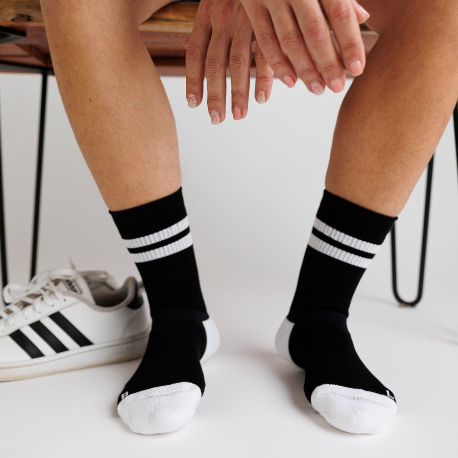 Seated person wearing black Retro crew grip socks beside white sneakers. By Squid Socks.