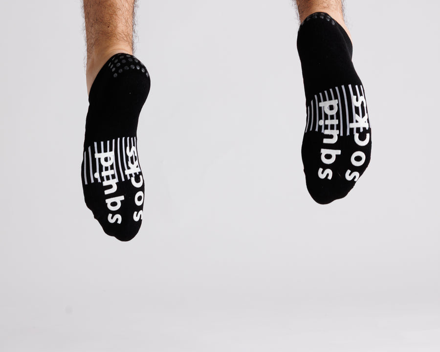 Black no-show grip socks with both soles facing the camera, showing white bottom grips. By Squid Socks.