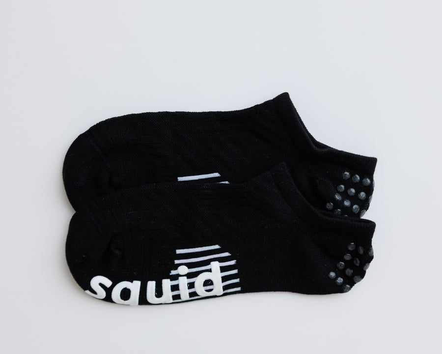 One pair of black no-show grip socks laid together, showing heel dots and white bottom grips. By Squid Socks.