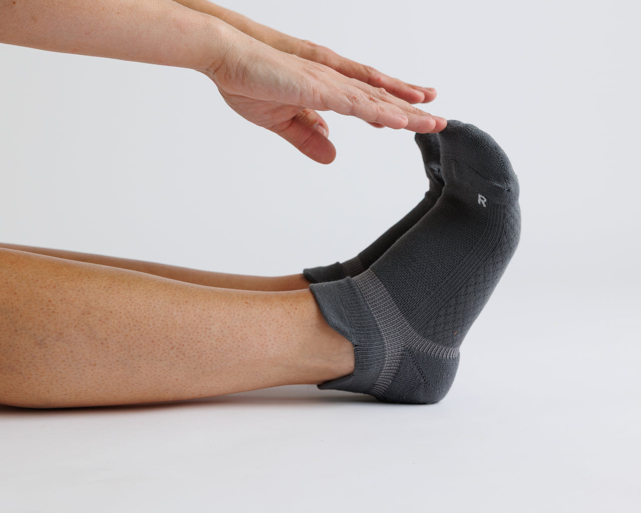 Person reaching toward a raised foot wearing a charcoal gray ankle grip sock. By Squid Socks.