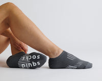 Thumbnail for Person seated in charcoal gray no-show grip socks, with one sole turned toward the camera. By Squid Socks.