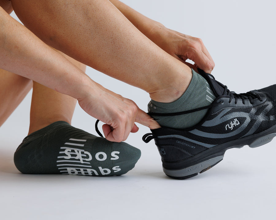 Person wearing muted green no-show grip socks while tying a black athletic shoe. By Squid Socks.