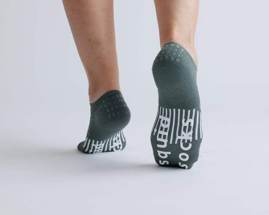 Muted green no-show grip socks with one heel raised, showing heel dots and white bottom grips. By Squid Socks.