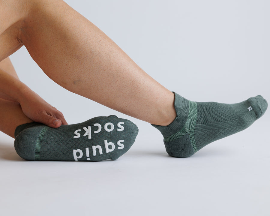 Person stretching a foot in muted green ankle grip socks, with white bottom grips visible. By Squid Socks.