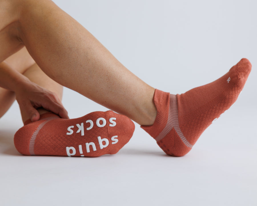 Person stretching a foot in terracotta ankle grip socks, with white bottom grips visible. By Squid Socks.