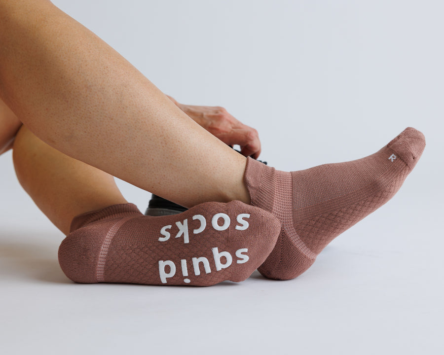 Person stretching a foot in brown ankle grip socks, with white bottom grips visible. By Squid Socks.