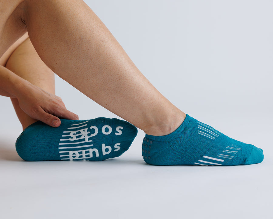 Person seated in teal no-show grip socks, with one sole turned toward the camera. By Squid Socks.