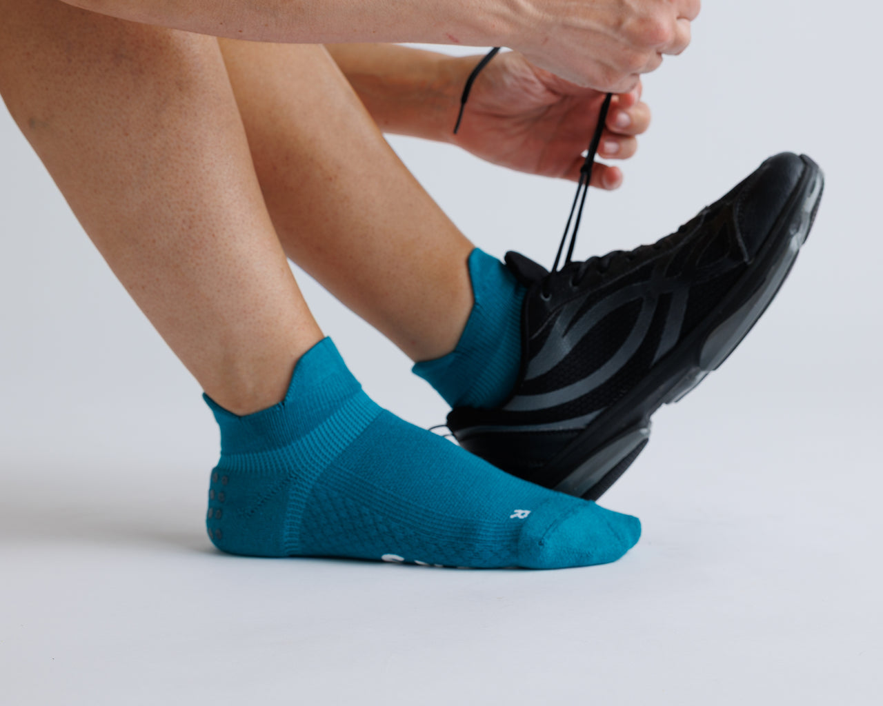 Person wearing teal ankle grip socks while tying a black athletic shoe. By Squid Socks.